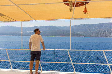 A man stands on the deck of a yacht and admires the sea and mountains on the horizon on a sunny day, relaxing and traveling.