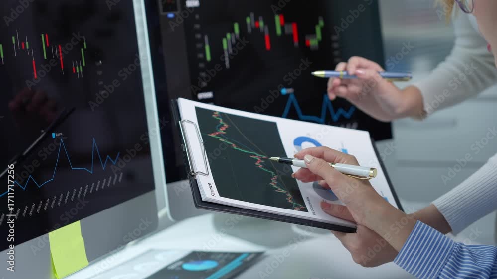Poster businesswomen discuss company performance, pointing at charts and graphs on a clipboard, with comput