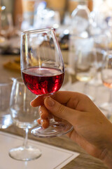 Close-up of a glass of rich red wine held by a man or a girl during a tasting. The background is blurred, you can see many glasses on the table, creating the atmosphere of a wine evening and enjoying
