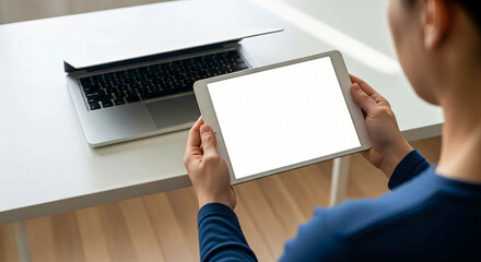 Over the shoulder view of a person holding a blank screen tablet in front of an open laptop on a white desk concept for remote work online learning or presentation