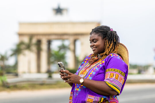 Smiling African woman in traditional purple dashiki uses smartphone outdoors for video calls, online shopping, digital banking, social media and internet browsing.