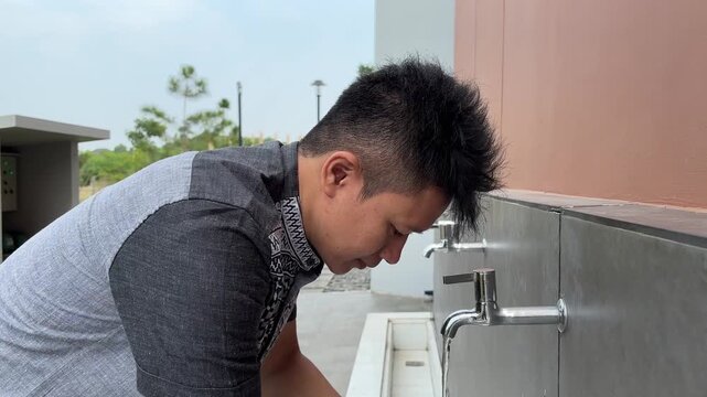 Muslim man rinsing mouth with clean water from faucet during ablution ritual before Islamic prayer outdoors
