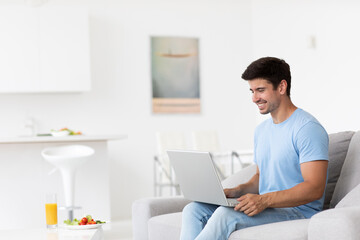 Cheerful guy is comfortably seated on a sofa in his own room with a laptop, smiling