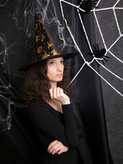 Beautiful girl witch in a witch's cap thoughtfully against the background of a web on Halloween