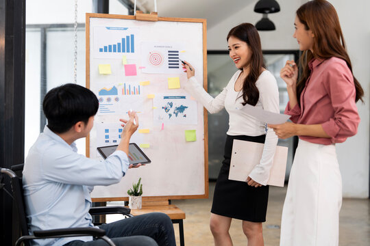 A diverse team of young professionals, including a man in a wheelchair, are collaborating on a new business project, using a whiteboard and a digital tablet to plan and analyze data in a office.