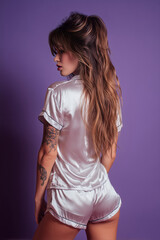 Elegant woman poses in silky sleepwear against a soft purple backdrop, showcasing body art and long wavy hair