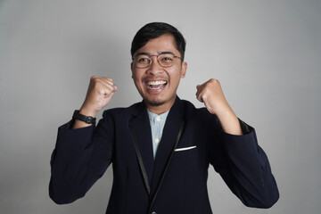 A happy Asian man in a blazer smiles with clenched fists raised in a gesture of celebration. This image is perfect for themes of business success, victory, and positive emotions.

