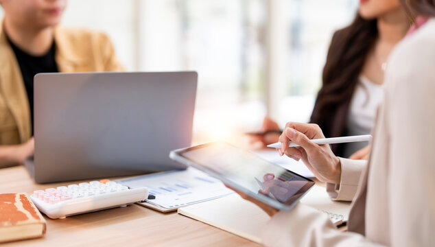 A close-up shot of a business team at a meeting, focusing on a woman using a digital tablet with a stylus. The scene highlights the use of modern technology for data analysis and collaboration. - Powered by Adobe