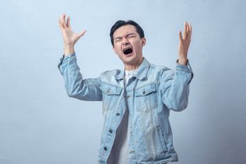 Naklejka premium Asian man in denim jacket screaming with both hands raised, expressing anger, frustration, sadness, or despair on a plain studio background, dramatic emotional reaction