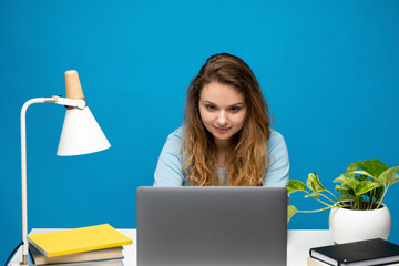 Young woman sitting at desk and working on laptop computer against blue background
