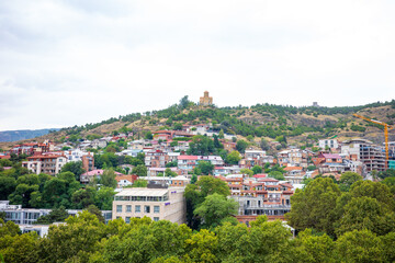 Fototapeta premium Colorful hillside neighborhood in Tbilisi, Georgia, with traditional houses, modern buildings, and a historic church on the hilltop. This scenic view captures the blend of old and new architecture