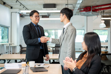 Asian businessman standing shaking hands and woman coworker clapping aside at wooden table in office