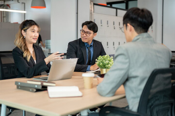 Businesswoman holding pen talking over laptop sitting with coworkers at wooden table in meeting room