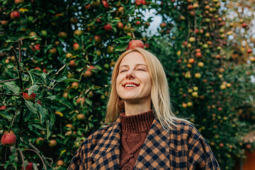 European woman stands by an apple tree in an autumn orchard, playfully balancing a ripe red apple on her head