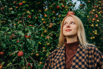 European woman stands by an apple tree in an autumn orchard, playfully balancing a ripe red apple on her head