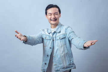 Asian man in denim jacket smiling with open arms gesture against plain background, symbolizing...