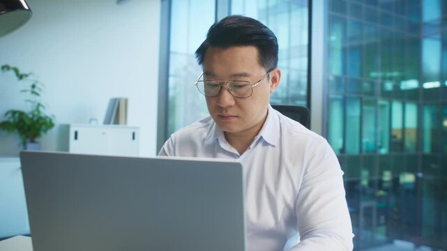 Serious Asian male in white shirt and glasses sitting at desk in bright office using laptop. Reading document or reviewing data. Concentrating on numbers or checking client presentation.