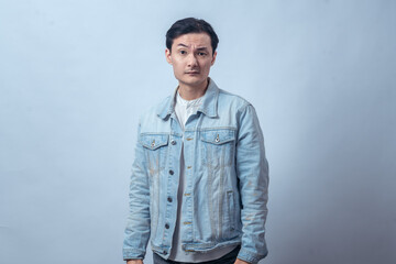 Fototapeta premium Asian man in denim jacket standing with hands on hips, raising eyebrow with doubtful expression, posing in studio against plain light background, showing skeptical and questioning attitude