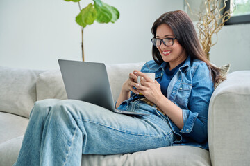 Young relaxed woman sitting on sofa at home with cup, looking at laptop