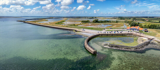 An aerial view captures a tranquil coastline with clear blue waters and a curved harbor in Zeeland, The Netherlands. A few cars near a restaurant are visible near the waterline.