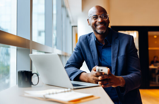 Smiling businessman using smartphone at a desk with laptop and notebook - Powered by Adobe