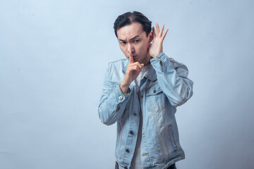 Fototapeta premium Asian man in denim jacket making silence gesture with finger on lips and hand cupped to ear, listening carefully, serious expression, concept of secret, hush, or confidential talk