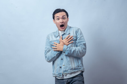Asian man in denim jacket showing scared expression, raising hands near face with open mouth in shock while standing against plain studio background