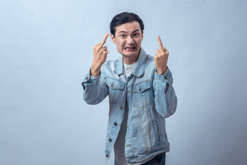 Asian man in denim jacket showing angry face and raising both middle fingers, standing in studio...
