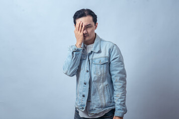 Fototapeta premium Asian man in denim jacket covering part of his face with one hand, looking serious and thoughtful, standing in studio against plain light background, showing stressed or worried expression