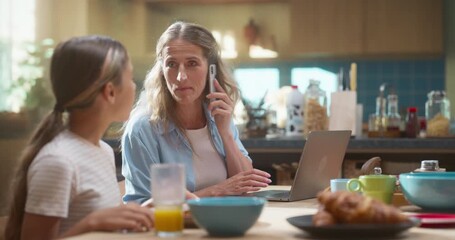Mother Efficiently Manages Remote Work on Her Laptop While Her Daughter Enjoys a Healthy Breakfast of Orange Juice, Cereal, and Croissants, Preparing for a Successful School Day