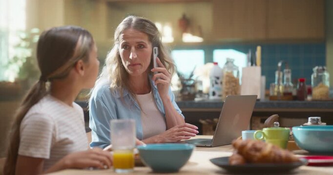 Mother Efficiently Manages Remote Work on Her Laptop While Her Daughter Enjoys a Healthy Breakfast of Orange Juice, Cereal, and Croissants, Preparing for a Successful School Day