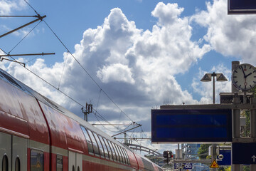 Modern Train Is At A Station With Overhead Lines A Clock And A Blank Blue Sign Under A Cloudy Sky