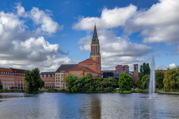 Fototapeta premium A Red Brick Church With A Tall Steeple Stands On The Shore Of A Lake With A Large Water Fountain