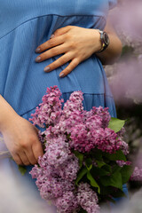 Fototapeta premium Pregnant woman in a blue dress gently cradles her belly while holding a bouquet of lilac flowers. Vertical photo, ideal for pregnancy, motherhood, floristry, family life and maternity photography