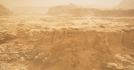 A large dust storm sweeps through a rugged desert terrain characterized by towering rock formations and a dramatic, hazy atmosphere in the late afternoon light.