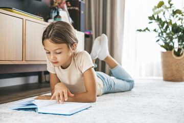 A young girl is lying comfortably on the floor, focused on reading a book. Education, childhood, and hobbies