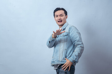 Asian man in denim jacket raising hand with serious face, making stop gesture while standing in studio against plain light background, showing warning sign and defensive expression