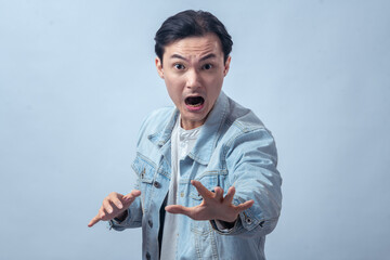 Asian man in denim jacket shouting with wide open mouth and raising hand forward, standing in studio against plain light background, expressing shock, anger, and strong emotional reaction