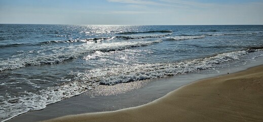 Sunny seascape with gentle waves breaking on a sandy beach, shimmering reflections of sunlight on the water, and a calm summer atmosphere.