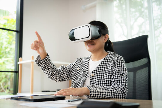 A woman wearing a black and white jacket is pointing at something while wearing a virtual reality headset. She is sitting at a desk with a laptop and a tablet