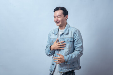 Asian man in denim jacket laughing hard, holding his chest with hand while closing eyes, showing...