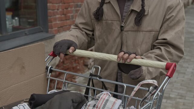 Midsection shot of unrecognisable mature male vagabond dressed in worn-out clothes pushing shopping cart with belongings while walking down city street