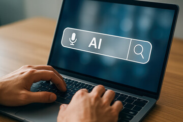 A person's hands typing on a laptop keyboard, with a search bar on the screen displaying 'AI' and a microphone icon.
