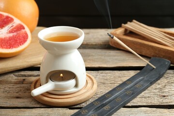 Aroma lamp with burning candle, smoldering incense stick and grapefruits on wooden table against dark background, closeup