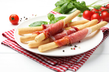 Delicious grissini sticks with prosciutto, peppercorns and basil on white wooden table, closeup