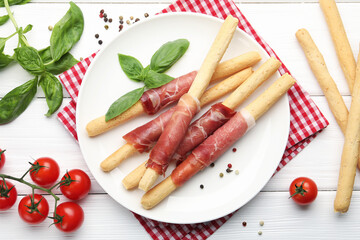 Delicious grissini sticks with prosciutto, peppercorns, tomatoes and basil on white wooden table, flat lay