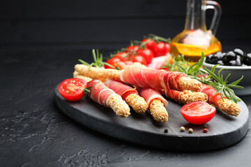 Delicious grissini sticks with sesame seeds, prosciutto, peppercorns and tomato on dark textured table against black background, closeup. Space for text
