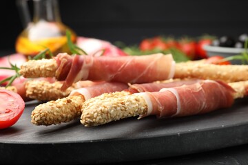Delicious grissini sticks with sesame seeds, prosciutto and tomato on dark table against black background, closeup