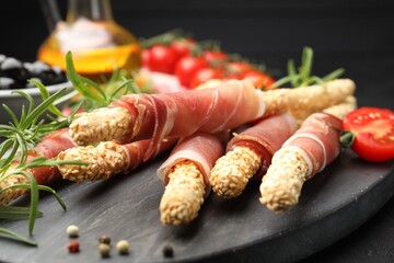 Delicious grissini sticks with sesame seeds, prosciutto, peppercorns, rosemary and tomato on dark table against black background, closeup