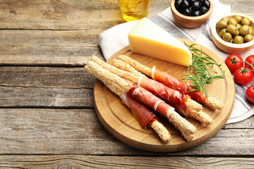 Delicious grissini sticks with sesame seeds, prosciutto and other products on wooden table, closeup. Space for text